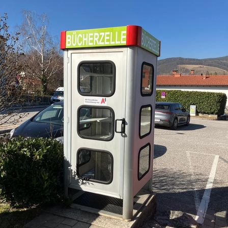 Ein Büchertauschkasten auf einem Bürgersteig mit einem grün-roten Schild, neben einem Parkplatz mit geparkten Autos und einem Gebäude mit rotem Dach im Hintergrund.