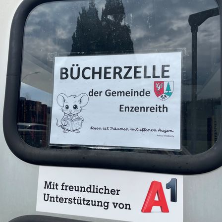 Ein Schild auf einer Busscheibe mit einer Maus, die ein Buch liest. Es steht 'Bücherzelle der Gemeinde Enzreith' und darunter befindet sich ein weiteres Schild, das 'Mit freundlicher Unterstützung von A1' lautet.