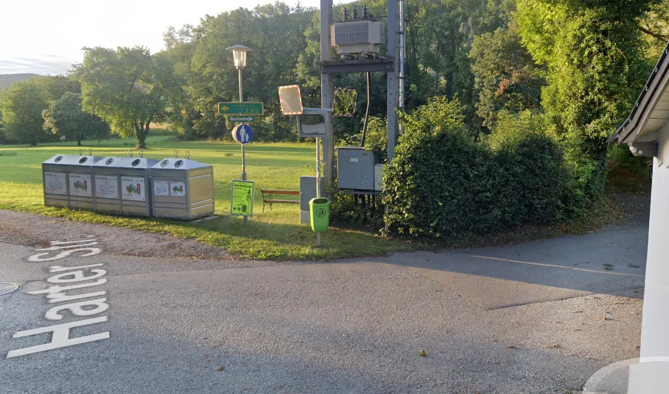 Ein Park mit Mülleimern, einem Abfallbehälter und einer Straßenlaterne ist zu sehen. Ein Schild mit einem Fußgängersymbol ist an einem Pfosten befestigt.
