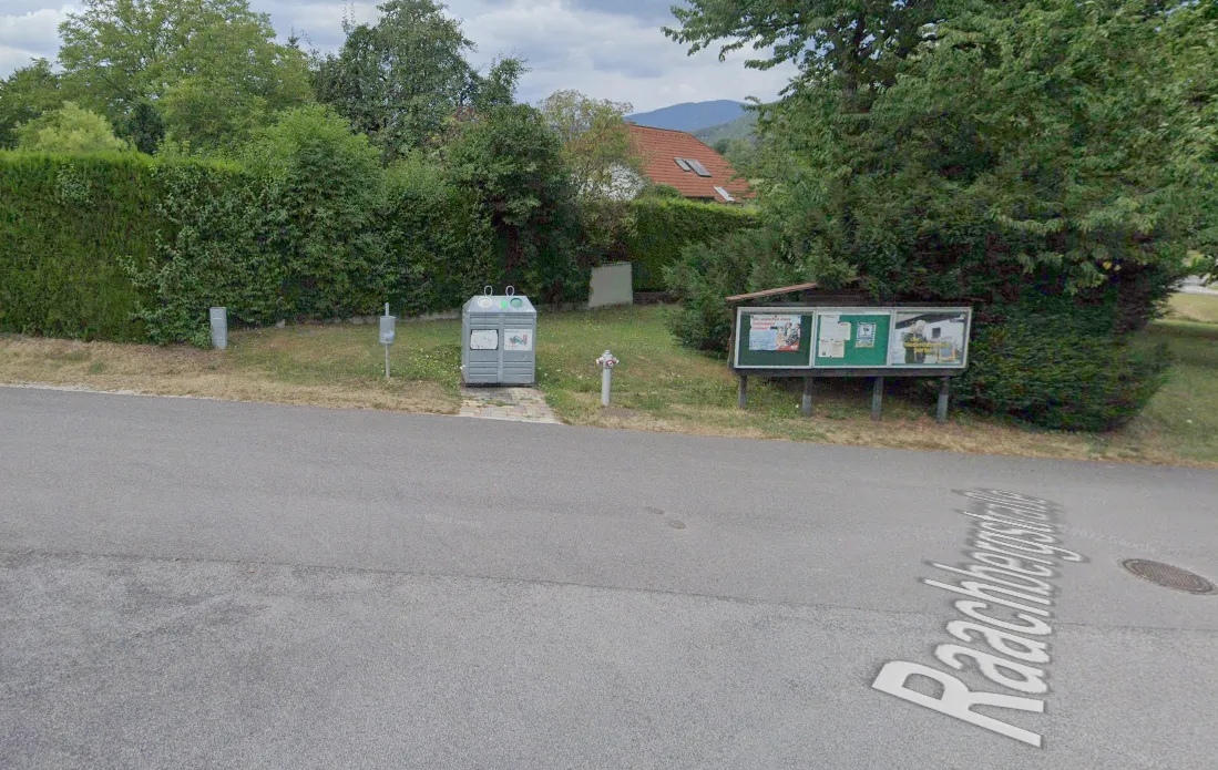 Eine Straße führt an einem Haus mit rotem Dach und einem Garten mit Bäumen und Büschen vorbei. Ein grauer Mülleimer steht neben einem Weg. Eine grüne Tafel ist am Boden befestigt.