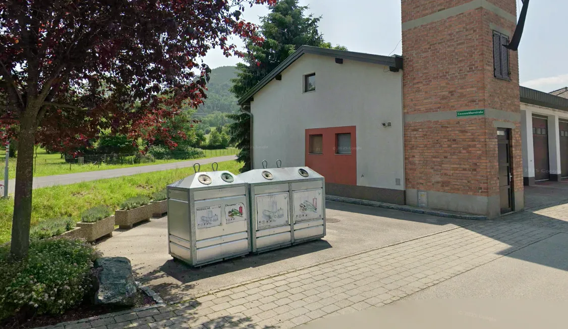 Drei Metallmüllbehälter stehen vor einem Haus mit Ziegelwänden und einem grünen Feld im Hintergrund.