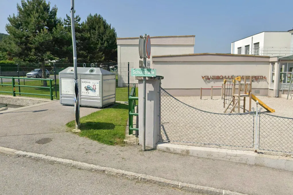 Eine Straßenansicht der Schußstraße mit einem grünen Mülleimer, einem Metallzaun und einem Gebäude mit einem Spielplatz im Hintergrund.