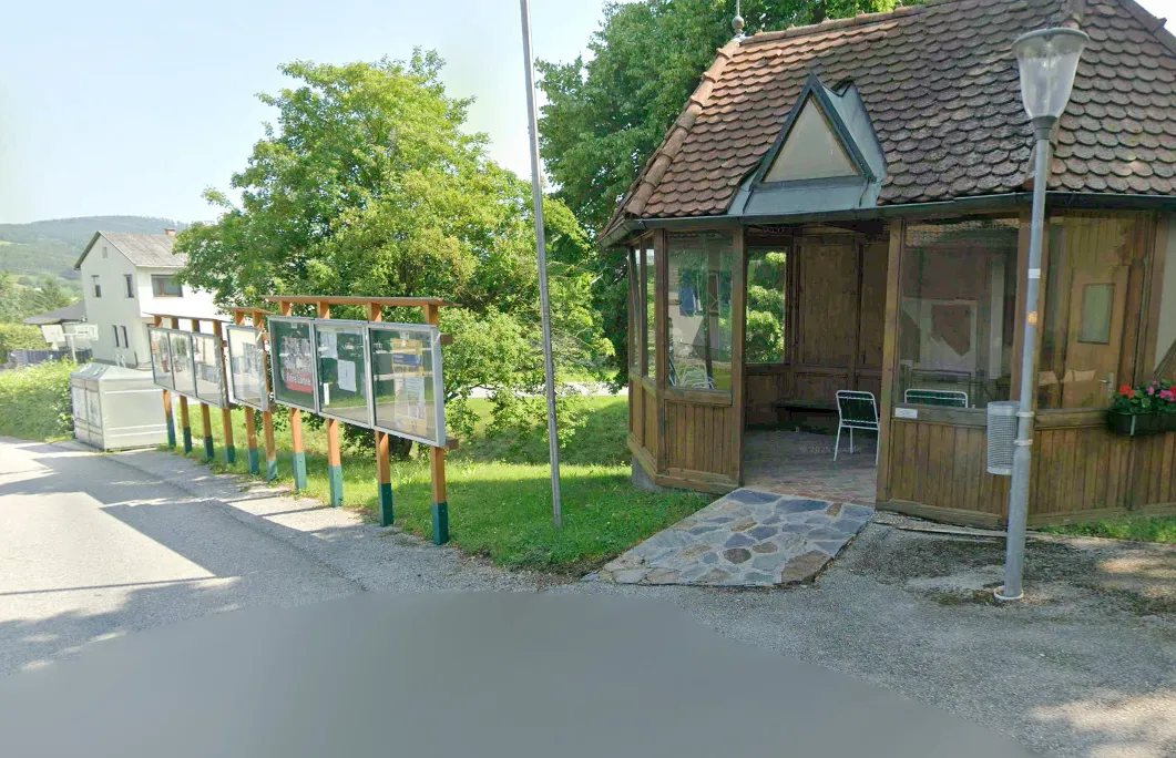 Eine kleine Holzhütte mit einem braunen Dach und Glaswänden befindet sich neben einer Straße mit einer Reihe von Informationsschildern. Die Hütte hat einen Steinweg, der zum Eingang führt. Bäume und Gras umgeben das Gebiet.