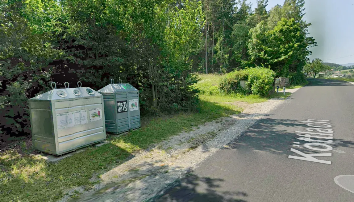 Zwei Mülleimer stehen am Straßenrand auf dem Rasen, einer hat einen Aufkleber für Recycling. Dahinter bilden dichtes Grün und ein Waldhintergrund eine ruhige ländliche Umgebung.