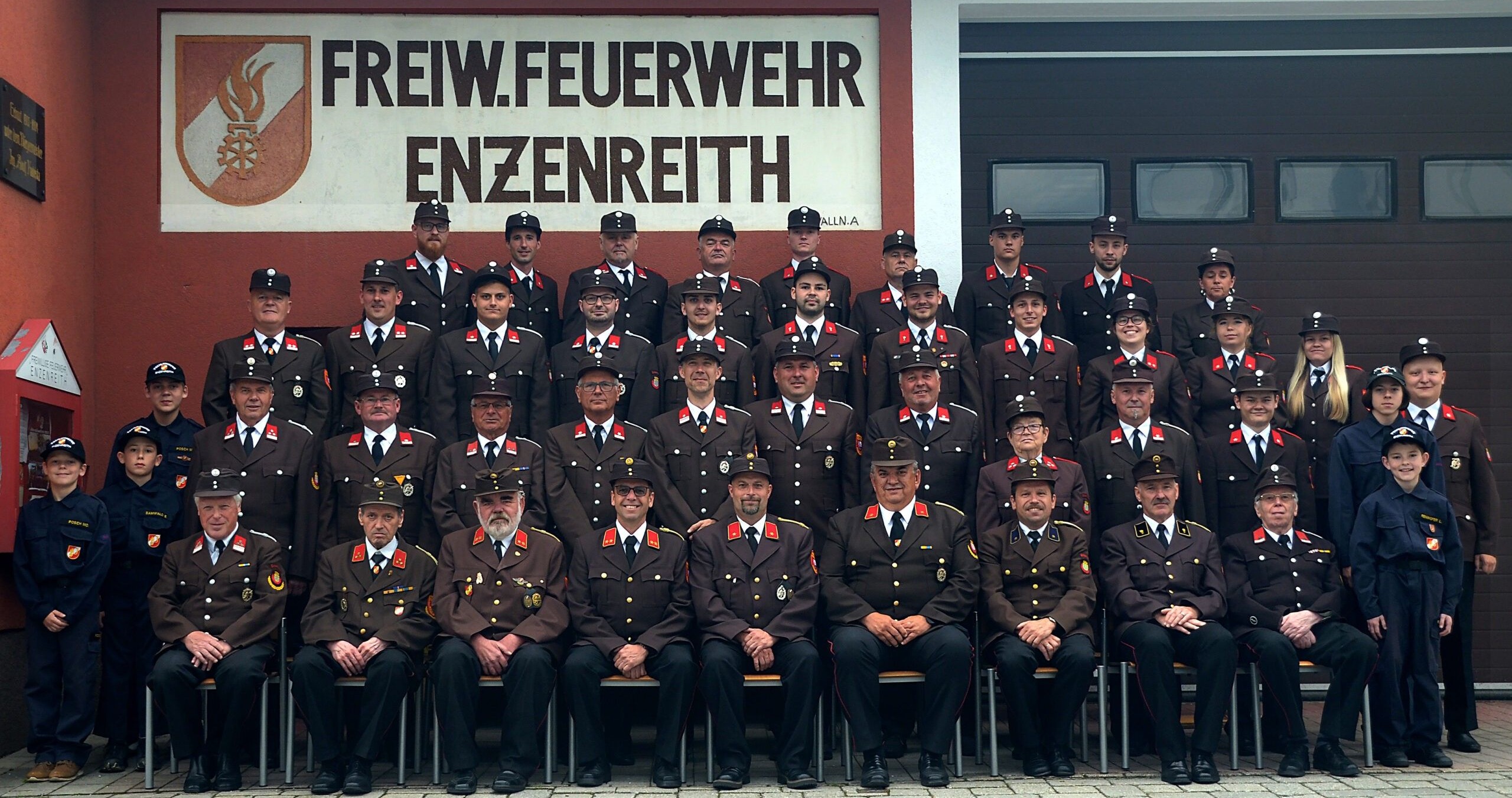 Eine Gruppe von Feuerwehrleuten in Uniform, stehend und sitzend, vor einem Gebäude mit einem Schild, das 'Reiw.Feuerwehr Enzenreith' steht. Sie alle schauen in die Kamera.