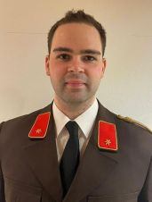 Ein Mann in einem Militäruniform steht vor einer weißen Wand und lächelt für ein Foto. Er hat rote Aufnäher mit Sternen auf seinen Schultern und Brust.