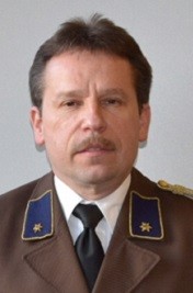 Ein Mann in Uniform, mit Schnurrbart und ernster Miene, steht vor einem schlichten Hintergrund.