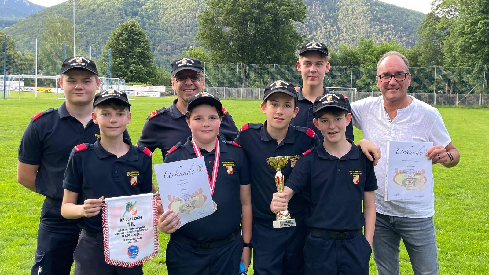 Eine Gruppe junger Menschen in Uniform posiert für ein Foto mit ihren Auszeichnungen. Einer hält einen Pokal, ein anderer ein Zertifikat. Hinter ihnen stehen lächelnde Erwachsene in Uniformen. Sie befinden sich auf einem Rasenfeld mit Bergen im Hintergrund.