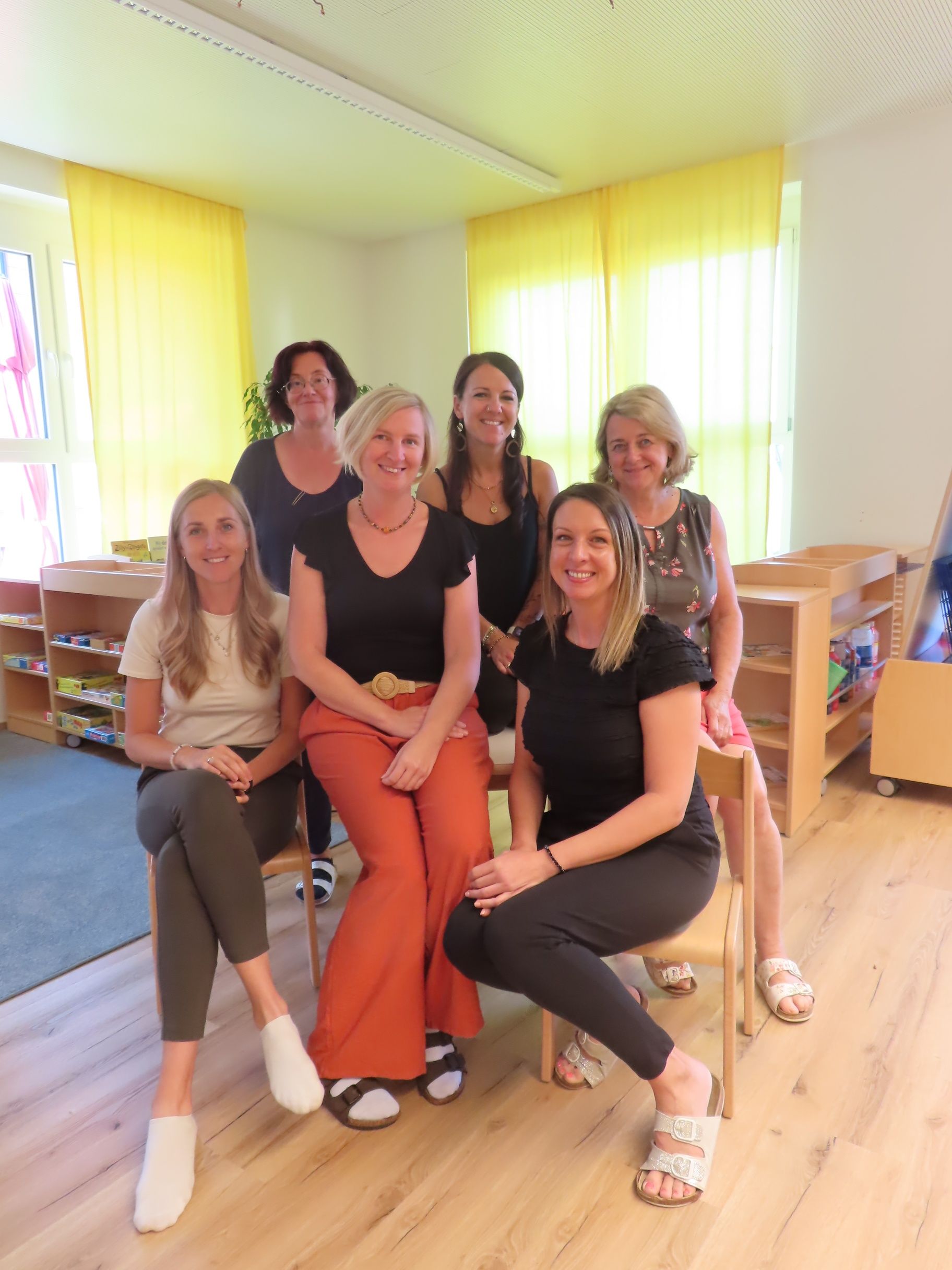 Sechs Frauen lächeln und posieren für ein Foto in einem Raum mit Holzböden und gelben Vorhängen.