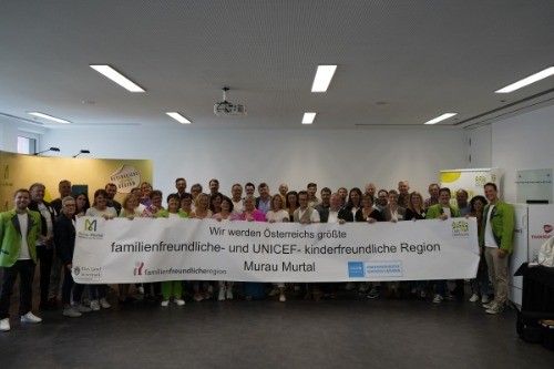 Eine Gruppe von Menschen, die ein großes Banner halten, steht in einem Raum. Sie tragen grüne Westen und Sneakers. Das Banner steht: Familienfreundliche und UNICEF-freundliche Region. Murau Murtal.