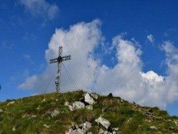 Ein Metallkreuz steht auf einem grasbewachsenen Hügel unter einem blauen Himmel mit Wolken. Das Kreuz ist an einem hohen Metallpfahl mit Drähten befestigt.