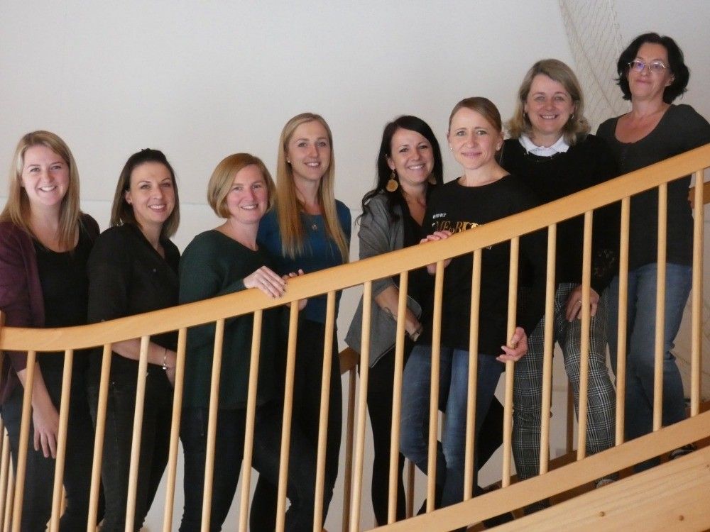 Sieben Frauen stehen auf einer Holztreppe. Sie lächeln und posieren für ein Foto. Sie tragen schwarze Kleidung. Die Frau rechts trägt eine karierte Hose. Die Holzgeländer der Treppe sind aus Holz.