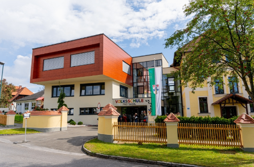 Die Volksschule Forstsaal, ein modernes Gebäude mit roter Fassade und Glasfenstern, ist an einem sonnigen Tag zu sehen. Menschen betreten das Gebäude und davor steht ein Zaun.
