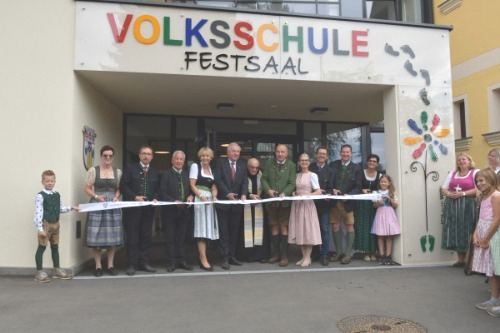Eine Gruppe von Erwachsenen und Kindern steht vor einem Gebäude. Sie halten ein Band, wahrscheinlich für eine Bandzeremonie. Das Gebäude heißt Volksschule Festsaal.
