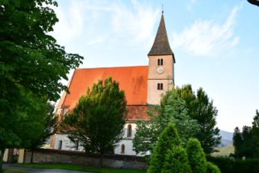 Eine traditionelle Kirche mit Turm und Uhr, umgeben von üppigen grünen Bäumen unter einem blauen Himmel.