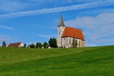 Eine kleine Kirche mit rotem Dach steht auf einem Hügel in einem ländlichen Gebiet, mit klarem blauem Himmel und verstreuten Wolken im Hintergrund.