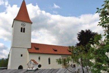 Eine weiße Kirche mit rotem Dach und einem Glockenturm, umgeben von grünen Bäumen und Büschen, unter einem bewölkten Himmel.
