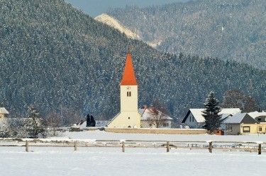 Eine kleine Kirche mit einem orangefarbenen Turm steht in einer verschneiten Landschaft. Dahinter befinden sich schneebedeckte Berge. Im Vordergrund sind mehrere Häuser und ein Zaun zu sehen.