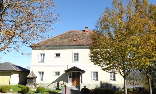 Ein weißes Haus mit einem braunen Dach und mehreren Fenstern befindet sich unter einem blauen Himmel. Vor dem Haus steht ein Baum mit gelben Blättern.