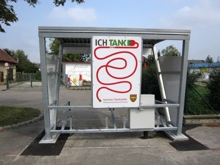 Eine metallische Struktur, die einer Bushaltestelle ähnelt, steht im Freien. Sie hat ein Schild mit 'ICH TANKE' und einem roten Spirallogo. Die Struktur steht auf einer gepflasterten Fläche mit Gras und einem Gebäude in der Ferne.