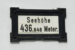 Ein schwarzer Rahmen an einer weißen Wand zeigt das Schild 'Seehöhe 436,648 Meter.' Es ist eine Höhenmessung über dem Meeresspiegel.