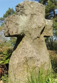 Ein verwittertes Steinkreuz steht in einem Garten, umgeben von Gras und Büschen, mit Bäumen im Hintergrund.