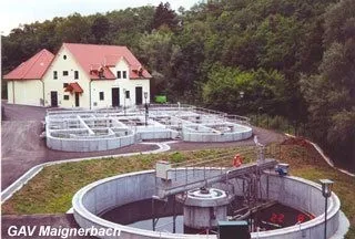Ein Luftbild einer Wasseraufbereitungsanlage mit einem rotgedeckten Haus und einem grünen Feld. Die Anlage verfügt über mehrere große runde Tanks und einen Wasserturm. Bäume umgeben das Gebiet.