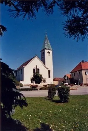 Eine kleine weiße Kirche mit Turm und Uhr steht auf einem Rasen, umgeben von Bäumen und Büschen.