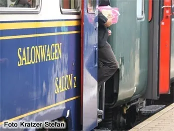 Eine Person mit einer rosa Tasche steigt in einen Zug mit blauen und gelben Akzenten, der mit 'Salonwagen' und 'Salon Z' beschriftet ist. Der Zug steht auf einem Bahnsteig.