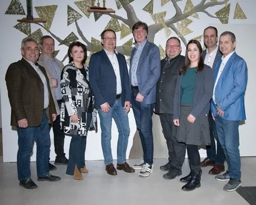 Eine Gruppe von Menschen in formeller Kleidung posiert zusammen vor einer Wand mit Baumdesign. Sie lächeln für ein Foto.