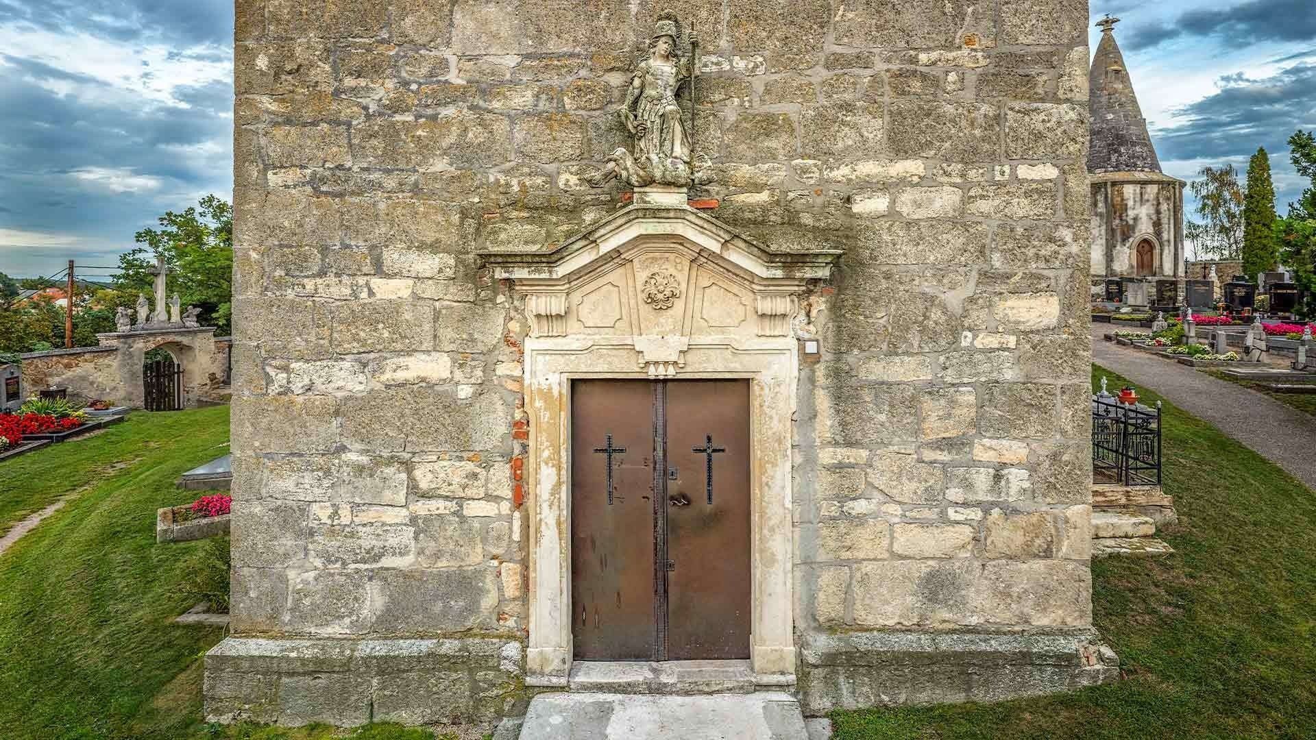 An einer alten Steinmauer befindet sich eine kleine Kapelle mit einer sitzenden Statue über einer Doppeltür. Die Tür hat zwei Kreuze und ein Löwenwappen über sich.