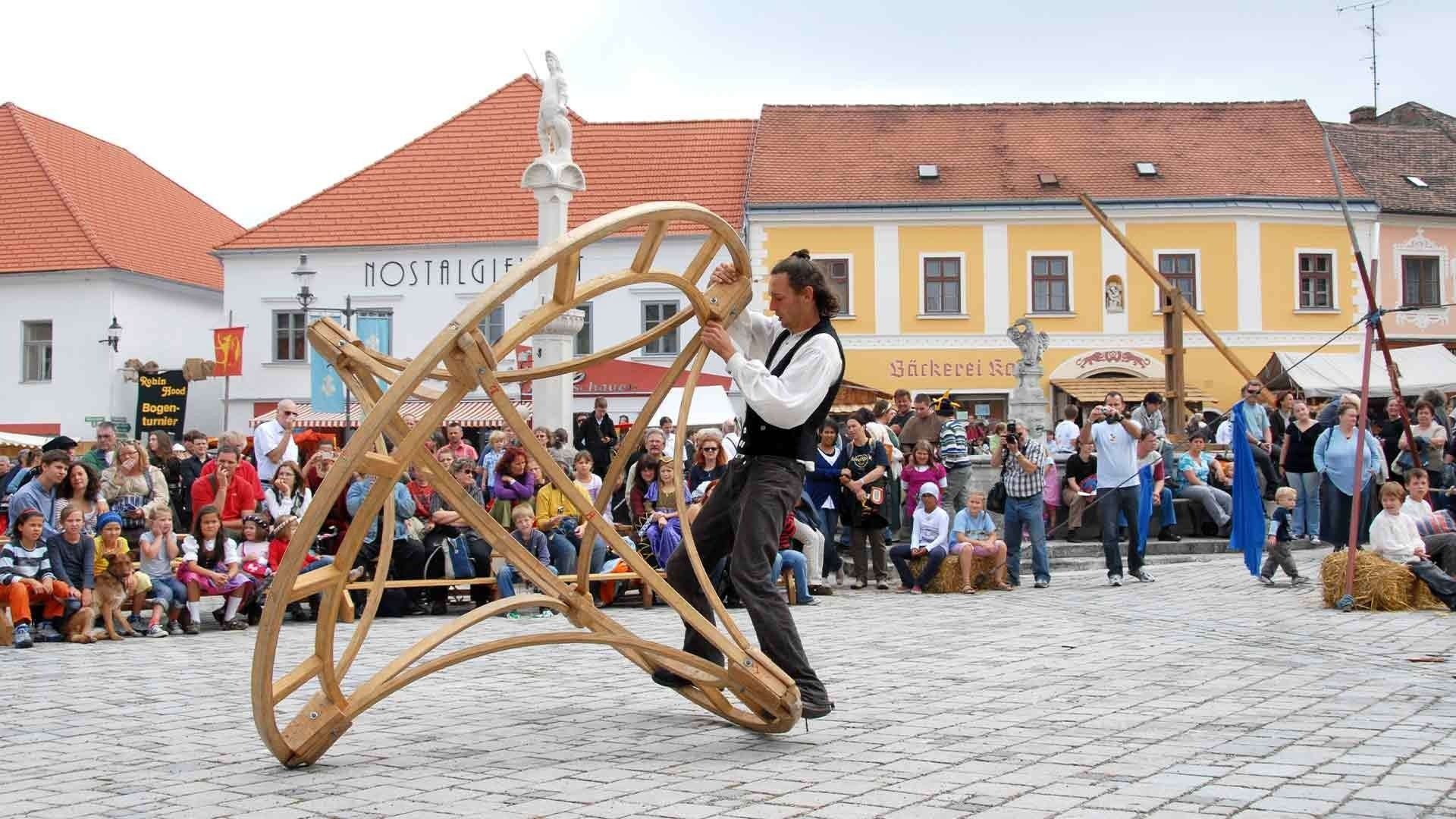 Ein Mann führt eine Holzskulptur auf einer Kopfsteinpflasterstraße vor einer Menge von Zuschauern vor. Eine Statue steht auf einem weißen Gebäude mit der Aufschrift 'Nostalgie'.