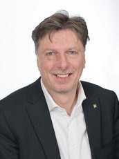 Ein Mann in einem schwarzen Anzug und weißem Hemd mit einem grünen Logo auf der linken Brust, er lächelt und zeigt die Zähne.