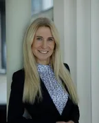 Eine Frau mit blonden Haaren und einem Blumenhemd lächelt vor einer weißen Wand.