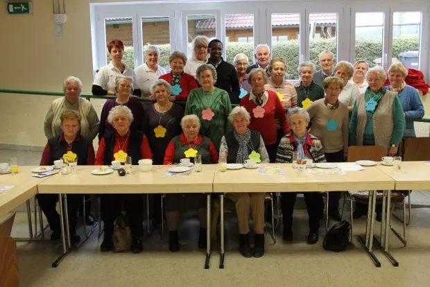 Eine Gruppe älterer Menschen versammelt sich um einen Tisch, einige sitzen und andere stehen. Sie lächeln und scheinen für ein Foto zu posieren. Der Tisch ist mit Tellern, Tassen und anderen Gegenständen gedeckt. Der Raum hat Glastüren und Fenster.