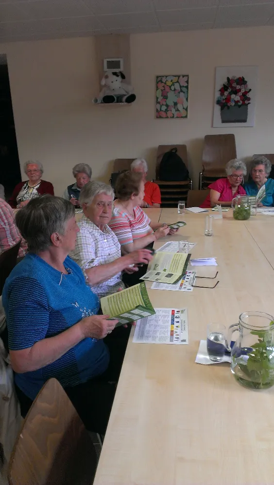 Eine Gruppe älterer Frauen sitzt um einen Tisch in einem Raum mit weißer Wand, einem Plüschtier und einigen Kunstwerken.