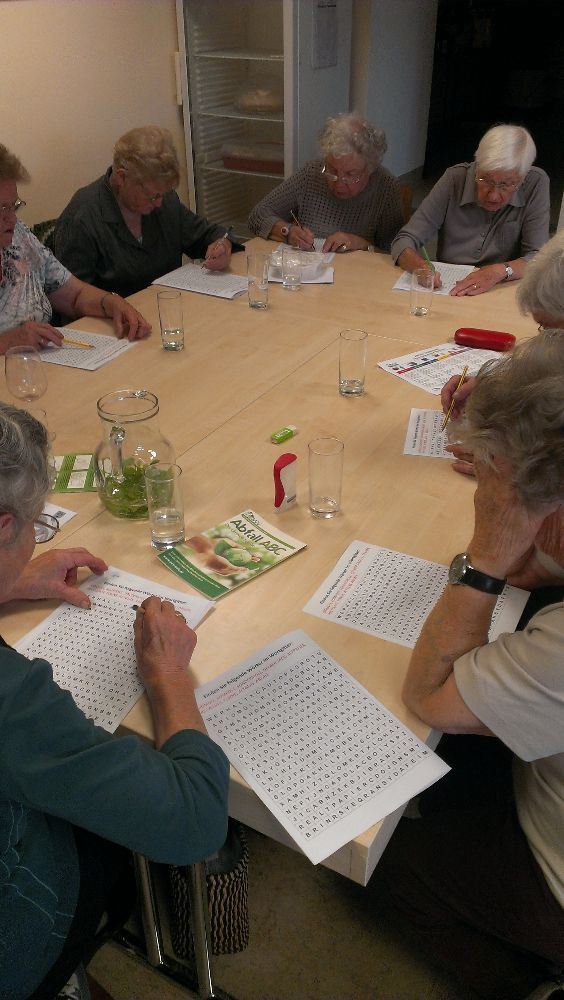 Eine Gruppe älterer Frauen sitzt um einen Tisch herum. Sie lösen ein Rätsel mit einem Stift.