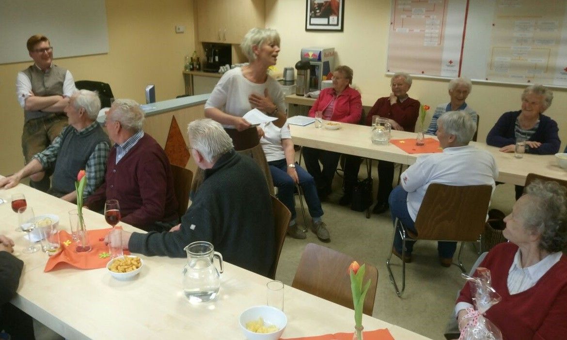 Eine Gruppe älterer Menschen sitzt in einem Raum um einen Tisch herum, lächelt und schaut sich gegenseitig an. Eine Frau steht und spricht mit ihnen.