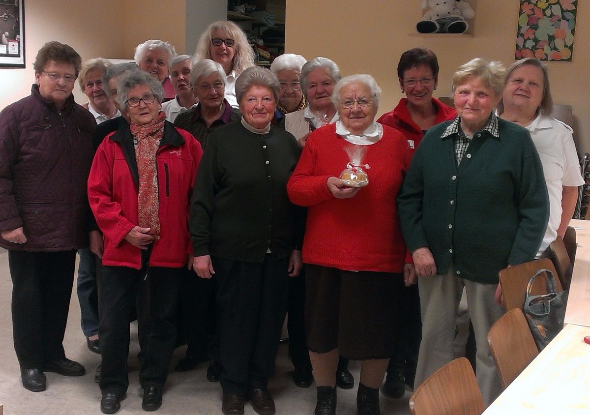 Eine Gruppe älterer Frauen und Männer steht in einem Raum, lächelt und posiert für ein Foto. Eine Frau hält einen Kuchen.