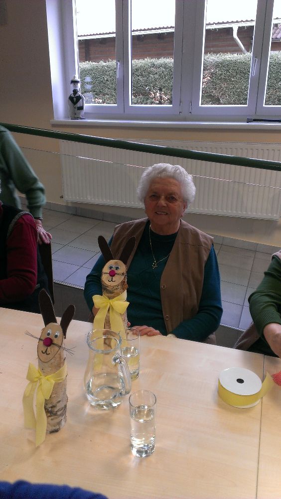 Eine ältere Frau sitzt an einem Tisch mit drei Osterhasen. Im Hintergrund ein weißer Heizkörper und ein Fenster mit einer Vase auf der Fensterbank.