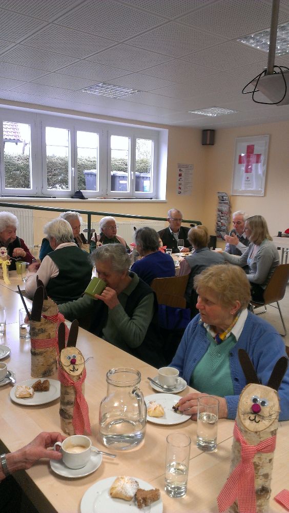 Eine Gruppe älterer Menschen sitzt an einem langen Tisch und unterhält sich. Jede Person hat eine Tasse und einen Teller vor sich, und einige trinken. Zwei Personen tragen Hasenkopfdekorationen.
