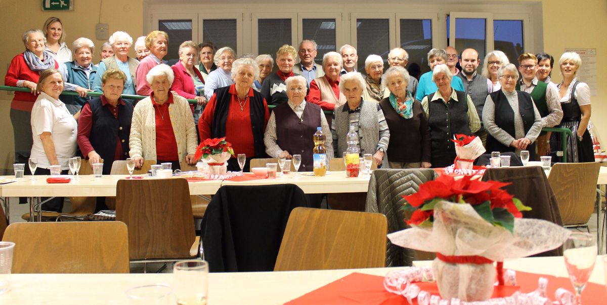Eine Gruppe von Menschen, sowohl Männer als auch Frauen, versammelt sich an einem langen Tisch für eine Weihnachtsfeier. Der Tisch ist mit Blumen und Weingläsern dekoriert.