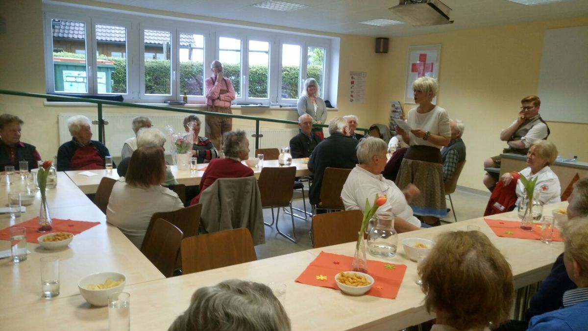 Eine Gruppe älterer Menschen versammelt sich in einem Raum, einige sitzen um Tische herum, andere stehen. Eine Frau liest von einem Papier. Der Raum hat große Fenster, und ein rotes Kreuz befindet sich an der Wand.