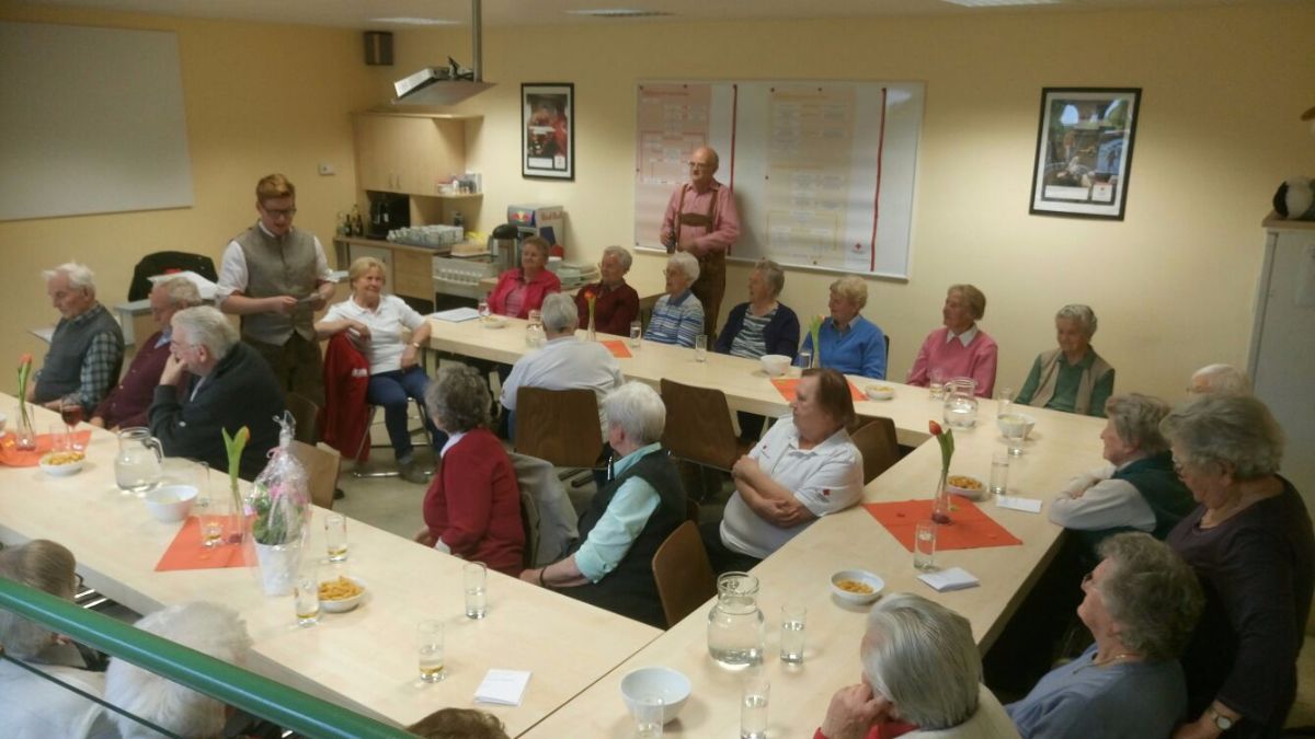 Eine Gruppe älterer Menschen sitzt um einen langen Tisch mit Essen, Wasser und Blumen. Ein Mann steht vor dem Tisch.