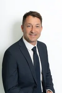 Ein Mann in einem dunklen Anzug und weißem Hemd, mit einer hellen Krawatte, lächelt und posiert für ein Foto vor einem schlichten weißen Hintergrund.