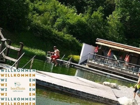 Ein Mann springt von einem Holzsteg in ein Gewässer. Ein Restaurant mit Terrasse und Sonnenschirmen befindet sich in der Nähe.