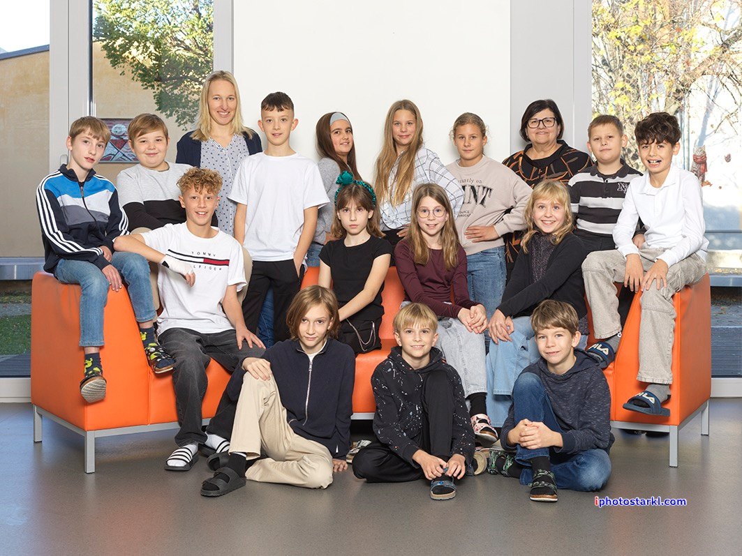 Eine Gruppe von Schülern und Lehrern posiert für ein Foto. Sie sitzen auf einer orangefarbenen Couch, einige stehen dahinter. Im Hintergrund ist ein großes Fenster zu sehen.