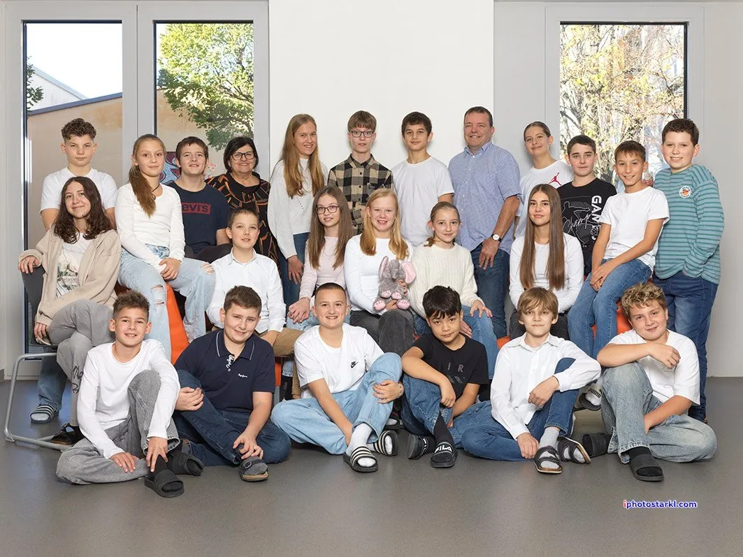 Eine Gruppe junger Menschen sitzt auf dem Boden in einem Raum. Sie tragen alle Freizeitkleidung. Einige sitzen auf einer Couch, während andere hinter ihnen stehen. Hinter ihnen befinden sich Glasfenster.