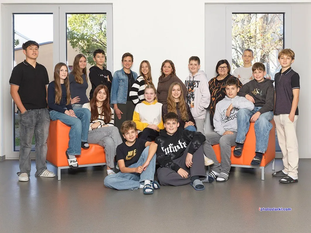 Eine Gruppe von Menschen sitzt auf orangefarbenen Stühlen in einem Raum. Einige tragen Brillen und Sneakers. Sie posieren lächelnd für ein Foto. Der Raum hat Glastüren und Fenster mit Bäumen draußen. Der Boden ist grau und hat einen Wasserzeichen in der unteren rechten Ecke.