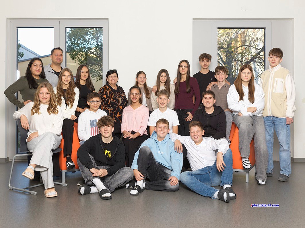 Eine Gruppe von Schülern und Lehrern posiert für ein Foto in einem Klassenzimmer, einige sitzen auf Stühlen und andere stehen dahinter. Der Raum ist gut beleuchtet durch natürliches Licht aus den Fenstern.